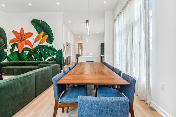 Bright modern open-plan dining room with long walnut table, blue upholstered chairs, green velvet sectional, large tropical wall mural featuring orange lilies, and sheer floor-to-ceiling curtains