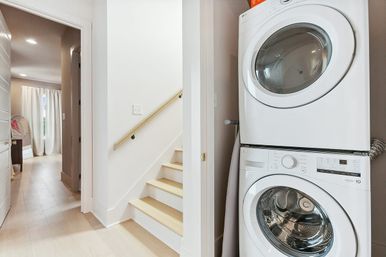 Sleek stacked washer and dryer in a bright hallway laundry nook beside light wood stairs and pale hardwood floors.