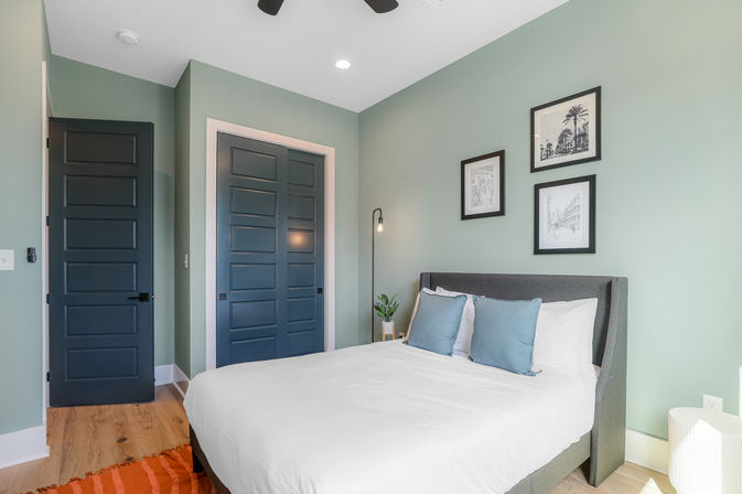 Cozy contemporary bedroom interior with mint-green walls, navy paneled doors, gray upholstered bed with white linens and blue throw pillows, framed black-and-white art, floor lamp and hardwood floors.