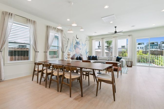 Bright modern open-plan dining and living room with a long wooden table and mid-century chairs, light wood floors, sectional sofa beneath an artistic mural, sheer curtains and sliding glass doors opening to a sunny fenced backyard with pool.