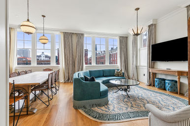 Bright open-plan urban living and dining room with large multi-pane windows and city view, teal curved sofa and glass coffee table on a patterned rug, long wooden dining table with woven chairs, hardwood floors, pendant lights and wall-mounted TV.