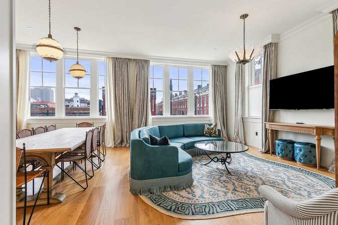 Bright open-plan urban living and dining room with large multi-pane windows and city view, teal curved sofa and glass coffee table on a patterned rug, long wooden dining table with woven chairs, hardwood floors, pendant lights and wall-mounted TV.