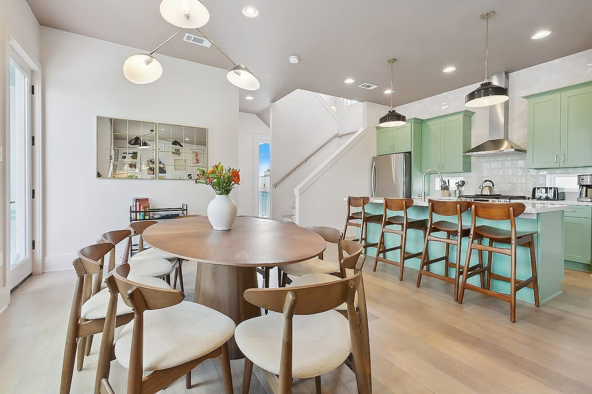 Bright, airy open-concept dining and kitchen with a round wooden table and cushioned mid-century chairs, mint-green island with four wooden bar stools, stainless appliances, modern pendant lights, hardwood floors, and a white vase of flowers.