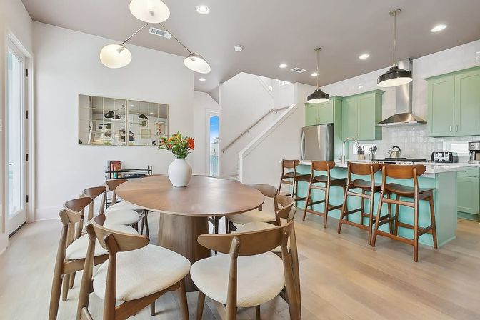 Bright, airy open-concept dining and kitchen with a round wooden table and cushioned mid-century chairs, mint-green island with four wooden bar stools, stainless appliances, modern pendant lights, hardwood floors, and a white vase of flowers.