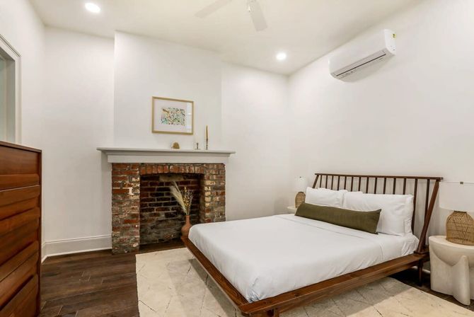 Cozy minimalist bedroom with a wooden platform bed and white bedding, exposed brick fireplace with decorative vase, dark hardwood floors, woven bedside lamps, and wall-mounted air conditioner.
