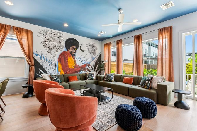 Bright modern living room in a sunlit apartment with a large colorful mural of a musician at a keyboard, blue painted ceiling with white fan, olive-green sectional, coral velvet swivel chairs, navy knitted poufs, rust-orange curtains and a black coffee table.