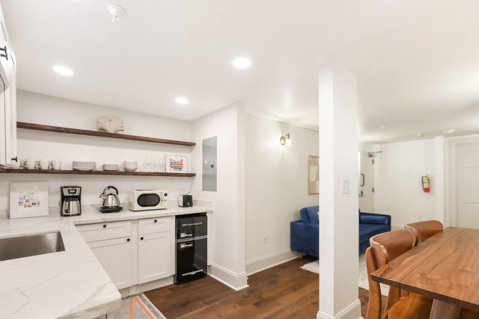 Cozy bright modern open-plan kitchenette and living area with white marble countertops, floating wood shelves, small black retro-style oven, coffee maker and microwave, blue velvet sofa, wooden dining table with leather chairs, and dark hardwood floors.