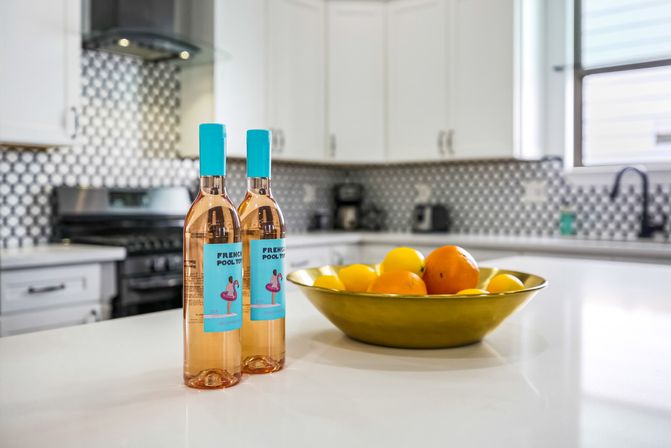 Bright modern white kitchen island with two rosé wine bottles featuring teal caps and labels beside a golden bowl of lemons and oranges, stainless stove and patterned tile backsplash in the background.