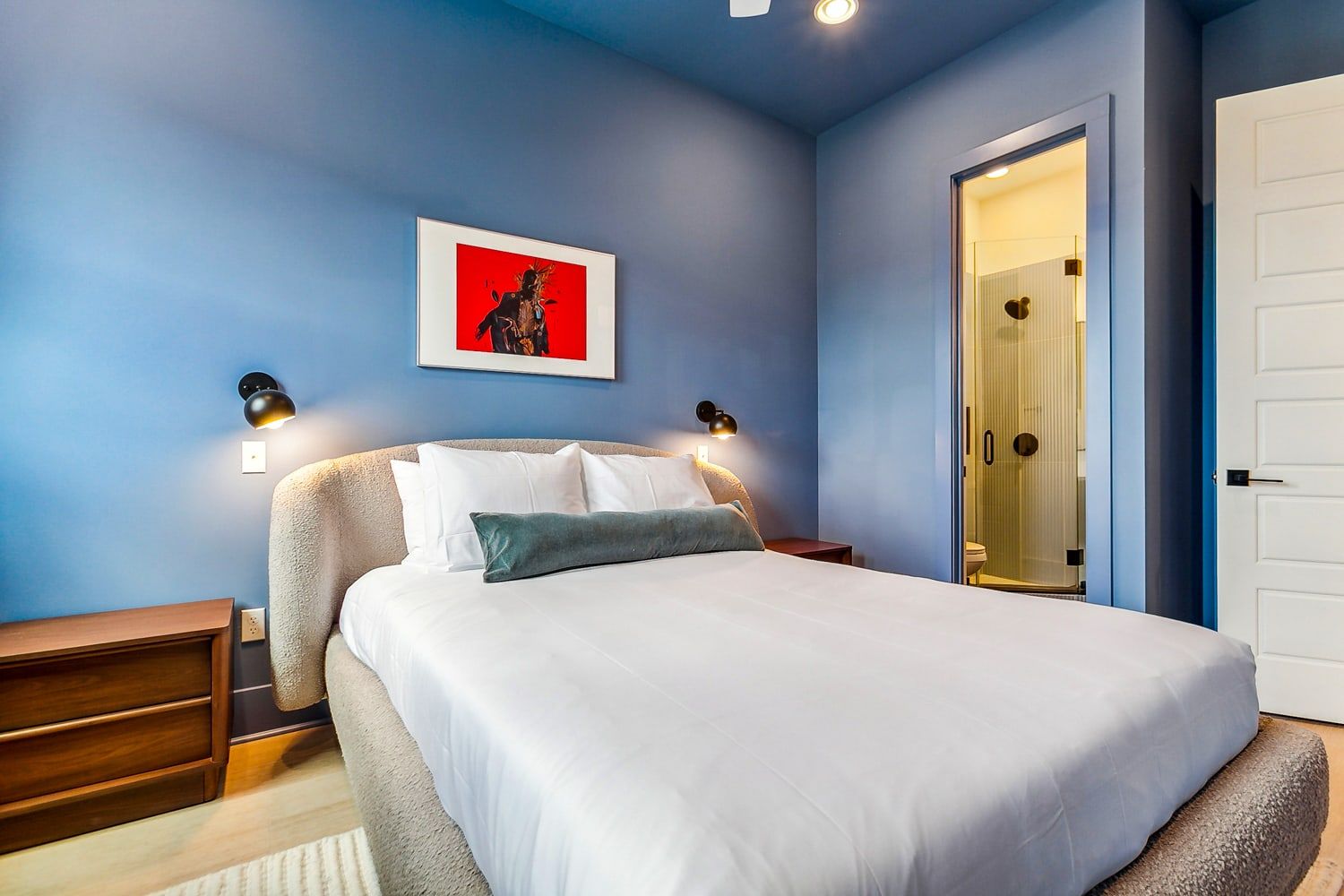 Cozy contemporary bedroom with blue walls, plush bed dressed in white linens and green bolster, wood nightstands, wall sconces, framed red artwork, and an en-suite glass shower visible through the doorway.