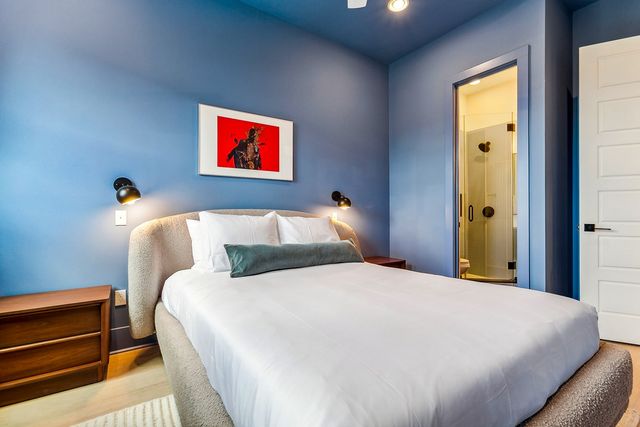 Cozy contemporary bedroom with blue walls, plush bed dressed in white linens and green bolster, wood nightstands, wall sconces, framed red artwork, and an en-suite glass shower visible through the doorway.