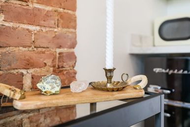 Rustic kitchen decor: wooden board on a black metal shelf in front of exposed brick, displaying two crystals and a brass candle holder with a tall twisted white candle.