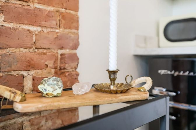 Rustic kitchen decor: wooden board on a black metal shelf in front of exposed brick, displaying two crystals and a brass candle holder with a tall twisted white candle.