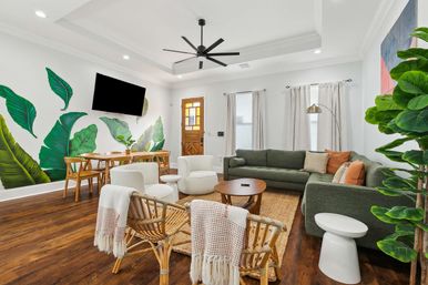 Bright modern open-plan living and dining room with tropical banana-leaf wall mural, green sectional sofa, rattan chairs with woven throws, round coffee table, wood dining set, wall-mounted TV, ceiling fan, hardwood floors and wooden front door
