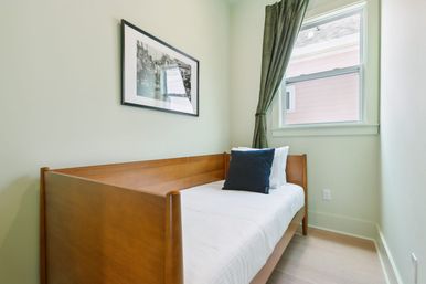 Cozy small guest bedroom with a wooden daybed, crisp white bedding and navy accent pillow, light-green walls, framed black-and-white photo, and a curtained window letting in natural light.
