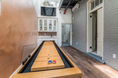 Loft-style game room with a wooden shuffleboard table ready for play, hardwood floors, gray exposed brick wall, copper-toned accent wall, glass-front cabinets and wall-mounted TV.