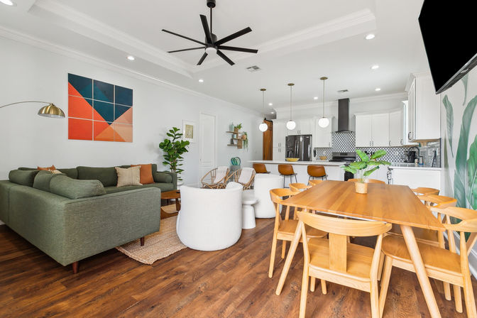 Bright open-concept living room and kitchen with a green sectional, cozy white lounge chairs, wooden dining table and chairs, white island with brown bar stools and globe pendant lights, geometric wall art, indoor plants, black ceiling fan and warm hardwood floors.
