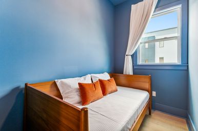 Cozy compact bedroom with blue walls and a wooden daybed dressed in white linens, two rust-orange accent pillows, and a sunlit window with a tied-back white curtain.