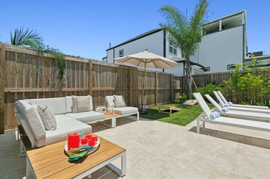 Sunny backyard patio with white sectional sofa and cushions, wooden coffee table with bright red drinks, umbrella and palm tree, artificial turf with cornhole boards, and four white sun loungers with rolled towels along a wooden fence.