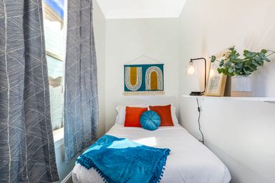 Cozy bright apartment bedroom with a single white bed, teal throw and round teal pillow, orange accent cushions, geometric wall hanging, gray patterned curtains by the window, and a floating shelf with a lamp and potted greenery.
