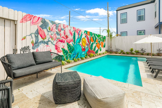 Residential backyard with a bright turquoise plunge pool, stone patio, gray cushioned sofa and poufs, black loungers and umbrella, and a colorful pink-and-green floral/tropical bird mural on a white fence.