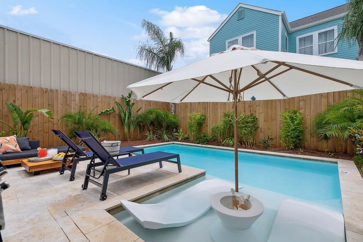 Sunny backyard plunge pool with built-in shallow loungers under a large white umbrella, two navy sunbeds on a stone patio, tropical plants and wooden privacy fence by a blue house.