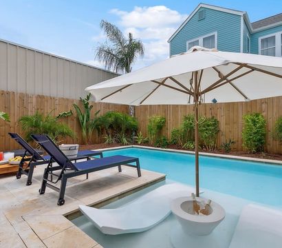 Sunny backyard plunge pool with built-in shallow loungers under a large white umbrella, two navy sunbeds on a stone patio, tropical plants and wooden privacy fence by a blue house.