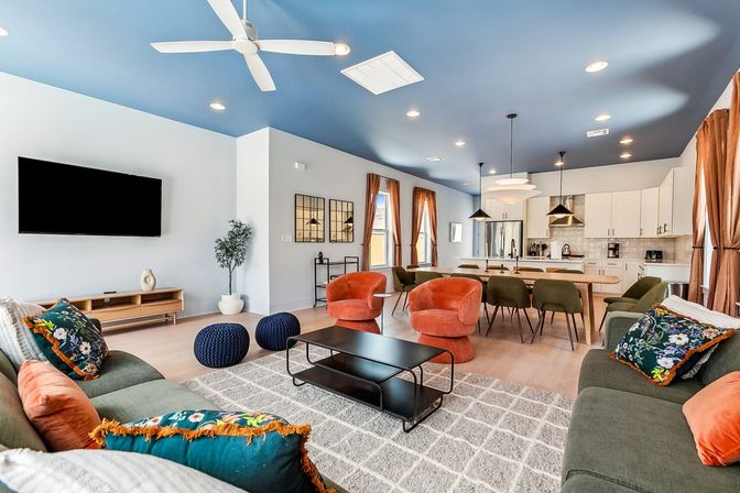 Vibrant open-concept living room and kitchen in a modern home with a bold blue ceiling, green sofas with colorful throw pillows, two orange swivel chairs, black metal coffee table on a gray rug, pendant lights over a long island and stainless steel appliances.