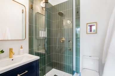 Sleek modern bathroom with glass-enclosed shower clad in vertical green tiles, brass rain showerhead and handle, navy vanity with brass faucet and wall mirror, and white toilet — compact, spa-like design.