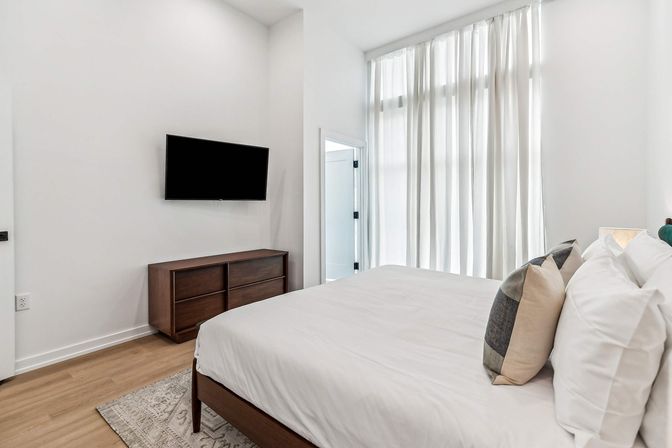 Bright modern minimalist bedroom with floor-to-ceiling sheer curtains, wall-mounted flat-screen TV above a walnut dresser, white bedding with accent pillows on a wooden bed frame, and light wood floors.