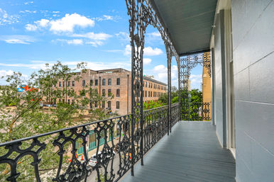 Sunny second-floor cast-iron balcony with ornate wrought-iron railing overlooking a tree-lined street and red-brick warehouse buildings in a historic downtown