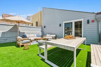 Sunny urban rooftop deck with bright artificial turf, wooden dining table and bench, cushioned lounge sofa set under a large umbrella and a tray of drinks on the table.