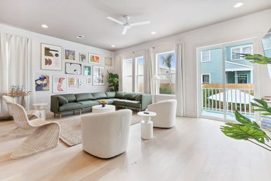 Sunlit modern living room with green sectional sofa, gallery wall of abstract art, white swivel chairs, light wood floors and sliding glass door opening to a balcony with a palm-tree view.