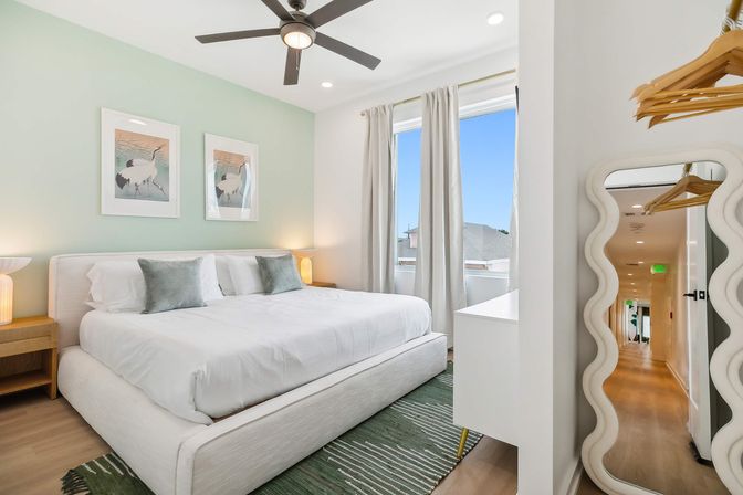 Sunlit modern bedroom with king-size bed, pale green accent wall, ceiling fan, two framed bird prints, bedside lamps, green rug, tall windows with curtains, and a wavy full-length mirror.