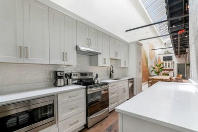 Sunlit modern open-concept loft kitchen with white shaker cabinets, quartz countertops, stainless-steel range and microwave, subway tile backsplash, skylight roof, and a dining table in the background.