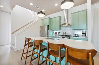 Bright modern open-concept kitchen with mint green cabinets, white quartz island and undermount sink, stainless steel fridge and range, four wooden mid-century bar stools, pendant lights and light wood floors.
