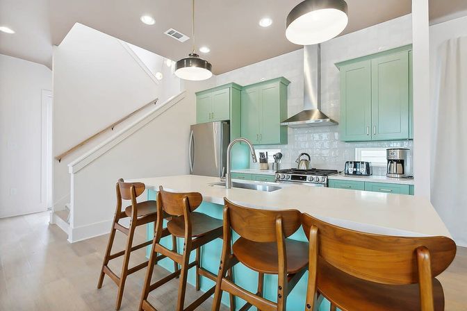 Bright modern open-concept kitchen with mint green cabinets, white quartz island and undermount sink, stainless steel fridge and range, four wooden mid-century bar stools, pendant lights and light wood floors.