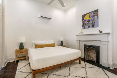 Cozy, bright minimalist bedroom with white walls, wooden mid-century bed frame and white bedding with mustard lumbar pillow, matching wood nightstands and lamps, decorative fireplace topped with abstract artwork, shag area rug and wall-mounted air conditioner.
