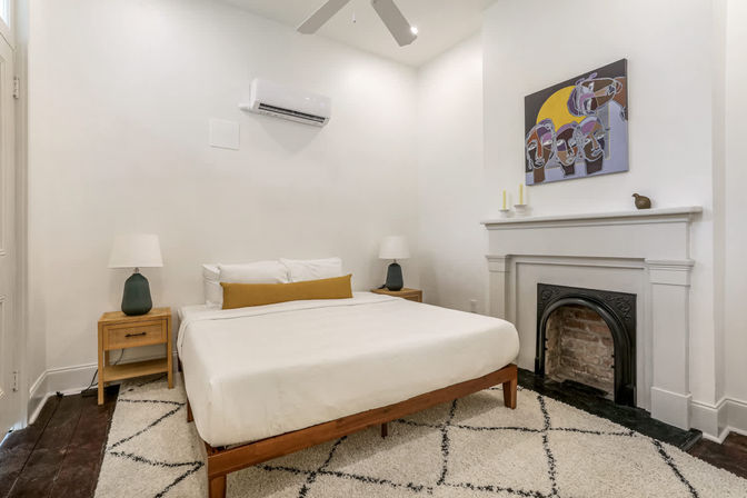 Cozy, bright minimalist bedroom with white walls, wooden mid-century bed frame and white bedding with mustard lumbar pillow, matching wood nightstands and lamps, decorative fireplace topped with abstract artwork, shag area rug and wall-mounted air conditioner.