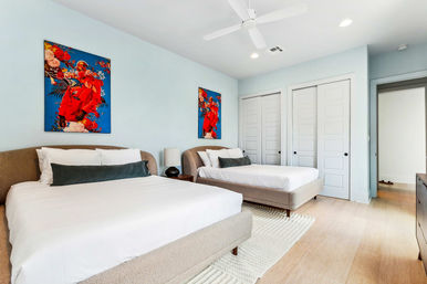 Bright modern bedroom with two upholstered beds and white bedding against pale blue walls, vibrant red floral artwork above each bed, ceiling fan, nightstand with lamp, double white closets, rug and light wood floors.