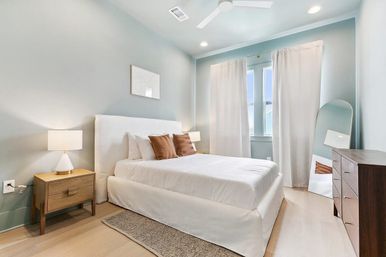 Sunlit modern bedroom with soft blue walls, white upholstered bed and bedding, two brown accent pillows, wooden nightstand with lamp, wooden dresser, full‑length leaning mirror, white curtains, ceiling fan, and light hardwood floor.