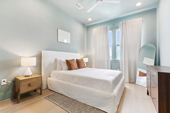 Sunlit modern bedroom with soft blue walls, white upholstered bed and bedding, two brown accent pillows, wooden nightstand with lamp, wooden dresser, full‑length leaning mirror, white curtains, ceiling fan, and light hardwood floor.