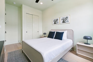 Bright modern minimalist bedroom with queen bed in white linens and navy pillows, light wood floors, matching bedside tables with black dome lamps, sliding closet doors and two framed black-and-white trumpet photos on a pale wall.