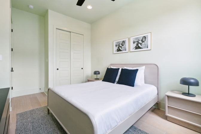 Bright modern minimalist bedroom with queen bed in white linens and navy pillows, light wood floors, matching bedside tables with black dome lamps, sliding closet doors and two framed black-and-white trumpet photos on a pale wall.