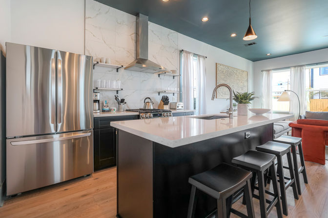 Sleek modern open-concept kitchen with large quartz island, stainless steel refrigerator and range, marble backsplash, black bar stools, pendant lights, and adjoining living area with a red sofa