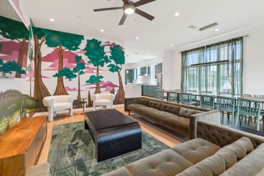 Bright open-concept living space with whimsical pink-and-green tree mural, tufted gray sectional sofas, two white accent chairs, black coffee table and green rug; adjoining dining area with long table, teal chairs and modern kitchen under a ceiling fan.