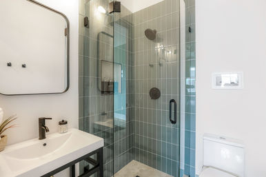 Sleek modern bathroom with glass-enclosed shower lined in vertical gray-green tiles, matte black shower fixtures, white vanity with black faucet and mirror, small plant on counter, and a white toilet.