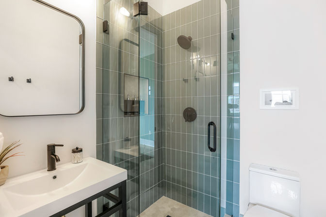 Sleek modern bathroom with glass-enclosed shower lined in vertical gray-green tiles, matte black shower fixtures, white vanity with black faucet and mirror, small plant on counter, and a white toilet.