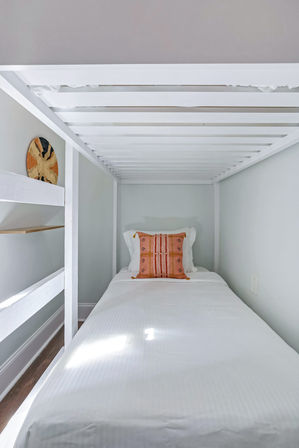 Minimal narrow bedroom with white wooden bunk frame and single bed dressed in crisp white linens, accented by a coral patterned throw pillow and woven wall basket — cozy small-space sleeping nook.