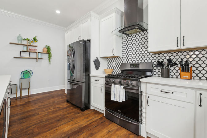Bright modern white kitchen with dark stainless appliances, black-and-white patterned tile backsplash, gas range and hood, white cabinets, warm hardwood floors, and floating shelves with plants.