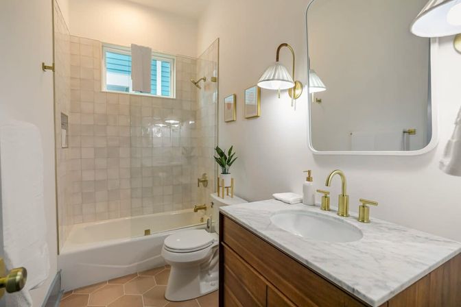 Bright modern bathroom with marble-top wood vanity, brass faucet and wall sconces, glass-enclosed bathtub-shower with white square tile, hexagon floor tiles, towel and small potted plant